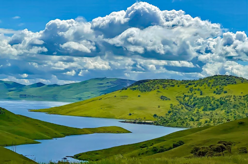 A meandering body of water amongst rolling green hills with fluffy clouds in a blue sky